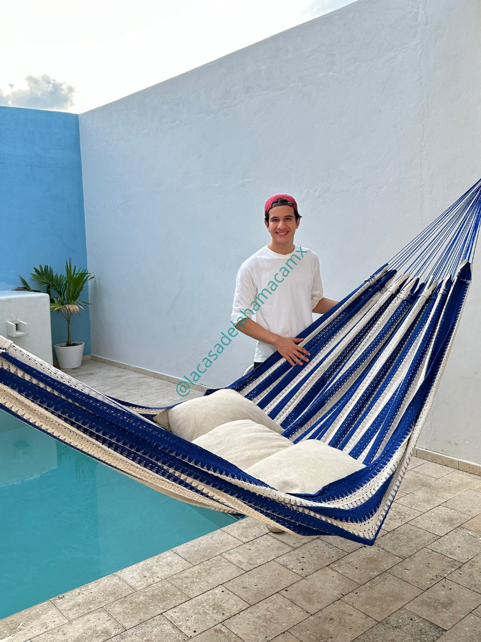 Hamaca mexicana azul y beige colocada en terraza junto a una alberca, con dos cojines y una persona sonriendo a un lado.