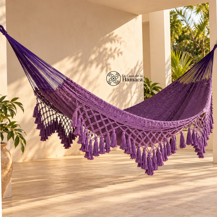 Hamaca yucateca artesanal de algodón morado con macramé colgada en una terraza con plantas tropicales
