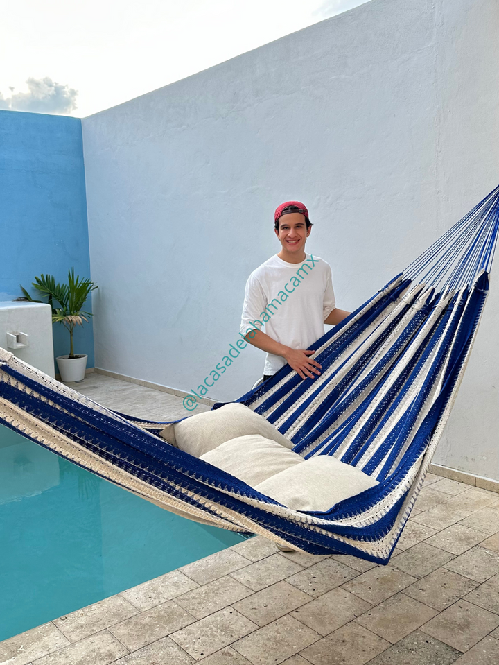 Hamaca mexicana azul y beige colocada en terraza junto a una alberca, con dos cojines y una persona sonriendo a un lado.