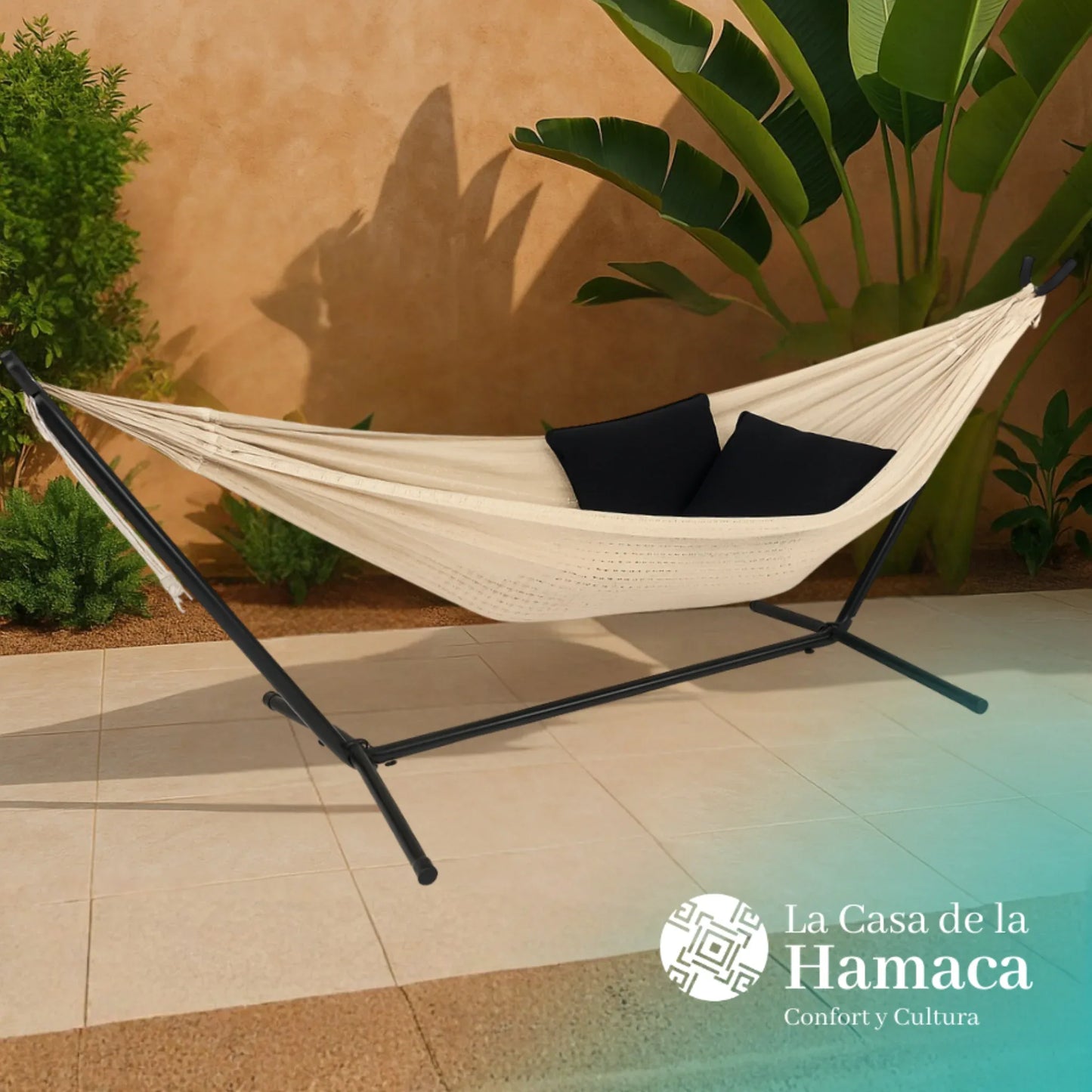 Hamaca mexicana beige de algodón colocada en base en exterior, con dos cojines negros y plantas de fondo
